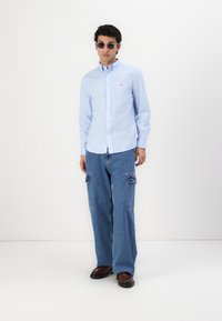 Camisa de botones de color azul claro, ajustada de forma relajada, con cuello. Pantalones cargo de mezclilla de pierna ancha con bolsillos laterales. Zapatos marrones y gafas de sol redondas.
