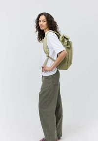 Zaino in tessuto verde con spalline regolabili; modello in camicia bianca e pantaloni olive larghi, in piedi contro uno sfondo semplice.
