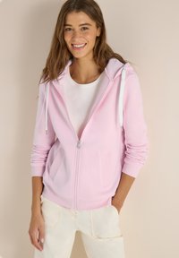 Jeune femme souriante, portant un sweat à capuche zippé rose clair sur un t-shirt blanc et un pantalon crème, debout devant un fond beige.