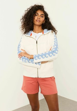 Woman with curly hair wearing white zip-up hoodie with blue wave patterns on sleeves and coral shorts, standing with arms crossed.