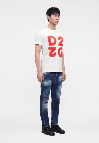Jeune homme debout portant un T-shirt blanc avec de grandes lettres rouges "DS2O", un jean bleu déchiré et des baskets noires sur fond blanc uni.