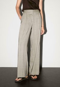 Pantaloni grigi chiari a gamba larga con un sottile motivo a scacchi, pieghe e una texture morbida; abbinati a flip-flop neri su una superficie scura.