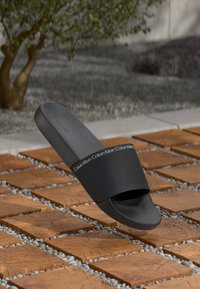 Fekete gumi csúszó szandál széles pánttal, fehér Calvin Klein logóval. Simább textúra, lapos talp és minimalista dizájn.