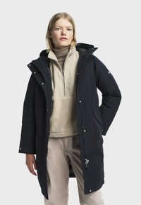 Femme blonde portant un pull à col roulé beige, un pantalon clair et un long manteau noir à capuche non fermé, devant un fond gris uni.