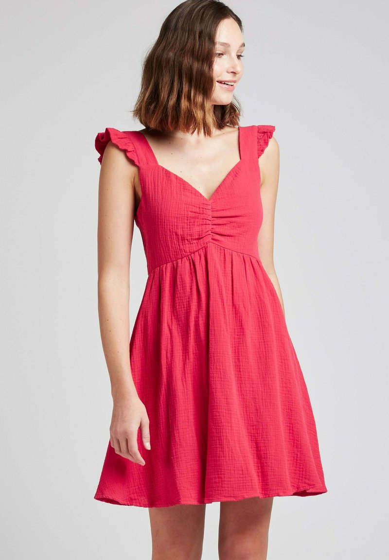 Robe en coton rose avec un corsage ajusté, des détails froncés et des bretelles à volants. Jupe évasée qui tombe au-dessus du genou.