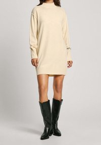 Abito maglione beige, lavorato a maglia, con colletto alto, polsini a coste e vestibilità ampia. Indossato con stivali alti in pelle nera.