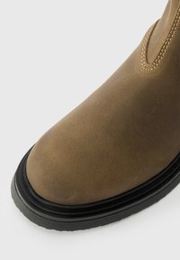 Καφέ δερμάτινη μπότες με λεία υφή, στρογγυλή μύτη και αντίθετη μαύρη καουτσούκ σόλα. Διαθέτει ραφή κατά μήκος της άκρης και του πλευρικού πίνακα.