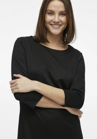 Svart långärmad topp i mjukt material med rund halsringning och avslappnad passform, visad på en modell med armarna i kors.