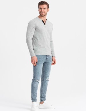 Mann trägt ein hellgraues Langarm-Henley-Shirt, ausgebeulte hellblaue Skinny-Jeans und weiße Sneakers und steht vor einem schlichten weißen Hintergrund.
