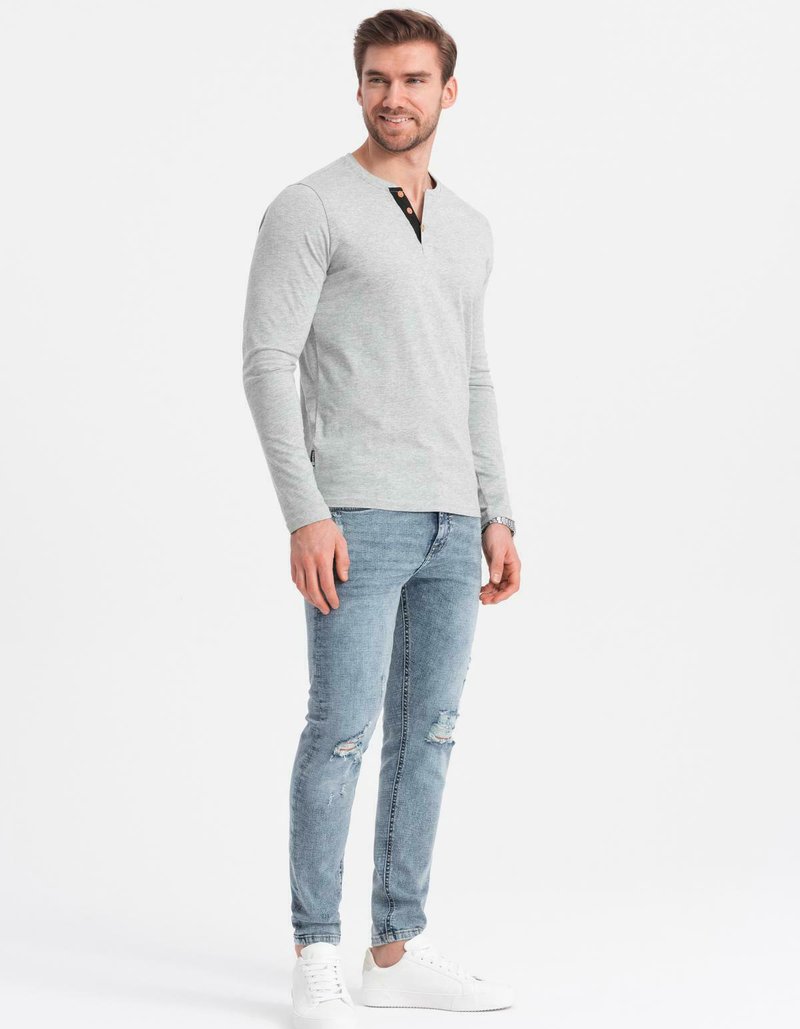 Man med ljusgrå långärmad henley-tröja, slitna ljusblå skinny jeans och vita sneakers, stående mot en enkel vit bakgrund.