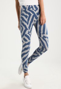Leggings a righe zebra blu e grigie, realizzati in tessuto morbido con una vita alta, abbinati a sneaker bianche su uno sfondo neutro.
