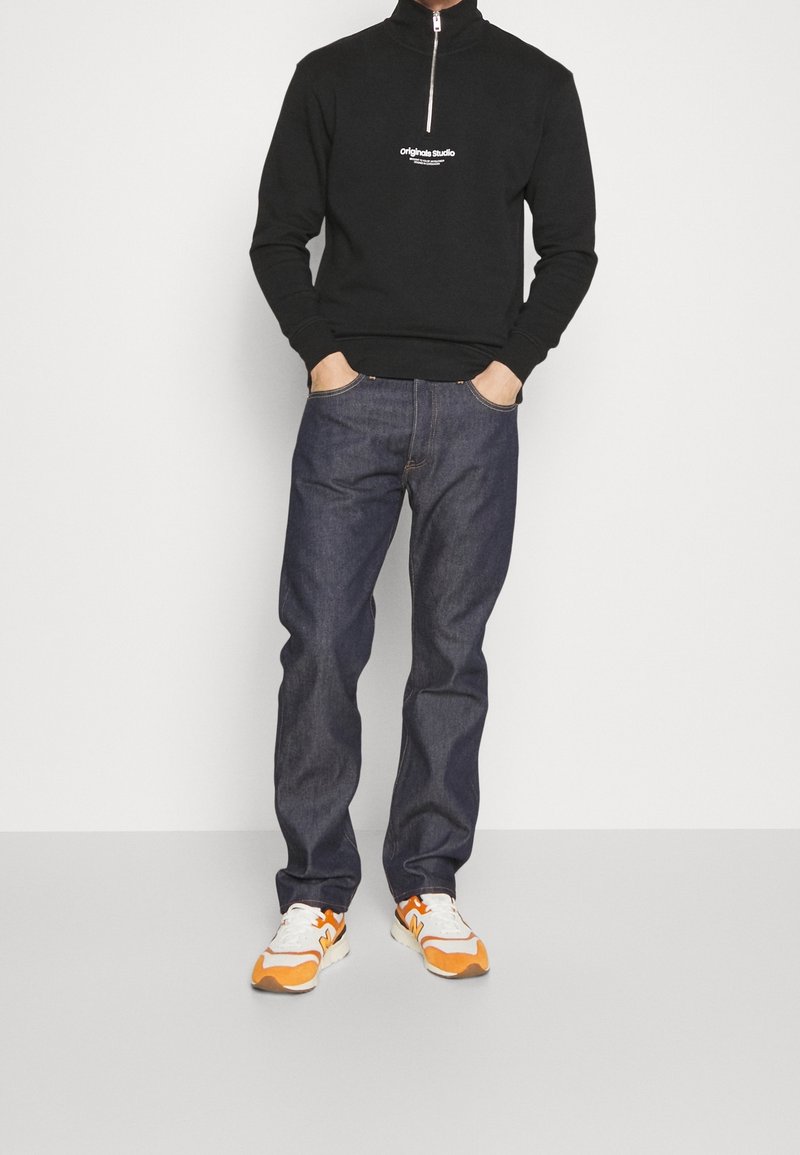 Sudadera negra con cremallera y logo en el pecho, combinada con jeans de mezclilla oscura. El calzado tiene una base blanca con acentos naranjas.