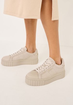 Osoba nosząca beżowe sneakersy na platformie w połączeniu z beżową spódnicą, stojąca na jasnej podłodze na neutralnym tle.