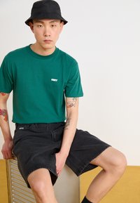 Green cotton t-shirt with white "OBEY" logo, paired with black denim shorts. Model sits on a metallic surface, wearing a black bucket hat.