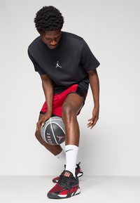 Svart kortermet t-skjorte med Jordan-logo, røde treningsshorts, hvite sokker og røde og svarte sneakers, holder en basketball.