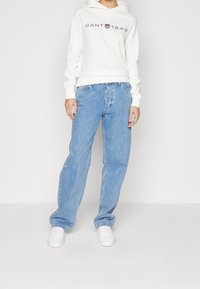 Sudadera blanca con el logo "GANT 1949", combinada con unos vaqueros de denim azul claro de corte holgado y zapatillas blancas, de pie contra un fondo liso.