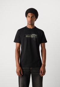 Tricou din bumbac negru cu mâneci scurte, având un imprimeu logo uzat în verde și alb pe piept, textură netedă, decolteu standard în formă de cerc.