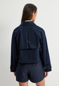 Giacca cropped blu navy con dettaglio a bavero sul retro, accenti di bottoni e colletto classico. Tessuto liscio con una vestibilità strutturata.