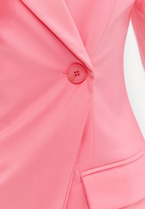 Gros plan d'un blazer rose montrant un seul bouton rose attaché au-dessus d'une poche à rabat sur le côté inférieur droit.