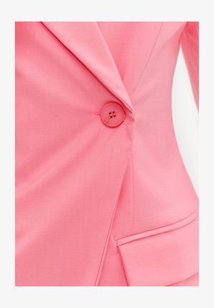 Gros plan d'un blazer rose montrant un seul bouton rose attaché au-dessus d'une poche à rabat sur le côté inférieur droit.