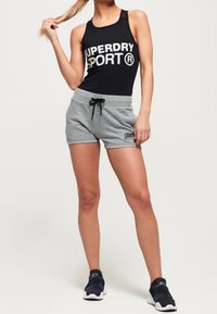 Camiseta sin mangas negra con el logo "SUPERDRY SPORT", combinada con pantalones cortos grises que cuentan con un cordón ajustable y un parche con el logo. Zapatillas deportivas completan el conjunto.