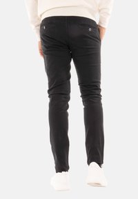 Pantaloni neri slim-fit in morbido tessuto, con due tasche posteriori dotate di chiusura a bottone e orli affusolati. Indossati con sneaker bianche.