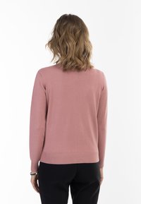 Maglione in rosa polveroso a maglia con scollo tondo, polsini e orlo a costine, e una texture liscia, indossato con pantaloni neri.