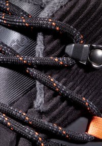 Tige de chaussure de sport noire en tissu texturé, avec des lacets tressés et des accents orange, ainsi qu'un mécanisme de verrouillage noir.