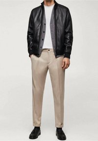 Zwart leren jasje met een grijze voering, gecombineerd met beige broek en zwarte schoenen. De outfit heeft eenjasje met knopen.