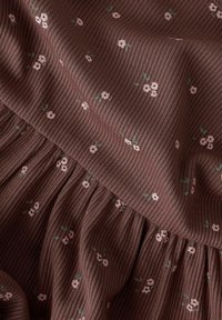 Tissu côtelé marron avec de petites fleurs rose pâle et des feuilles vertes, avec un design froncé à volants en bas.