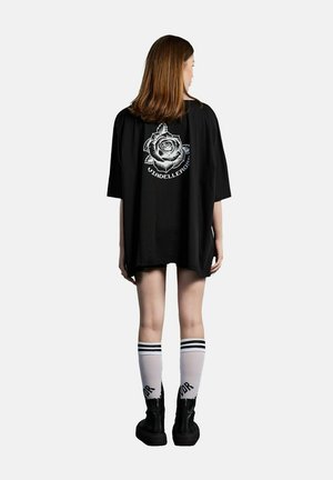 Femme debout, dos à la caméra, portant un t-shirt noir ample avec une rose blanche et un texte au dos, des chaussettes blanches à rayures noires et des bottes noires.