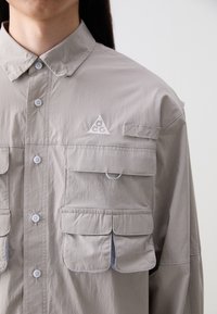 Light gray button-up shirt made of lightweight fabric, featuring multiple front pockets, a triangular logo, and a structured collar.