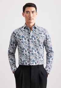 Uomo in camicia bianca a maniche lunghe con stampa floreale blu, pantaloni neri, in piedi con le mani in tasca contro uno sfondo semplice.