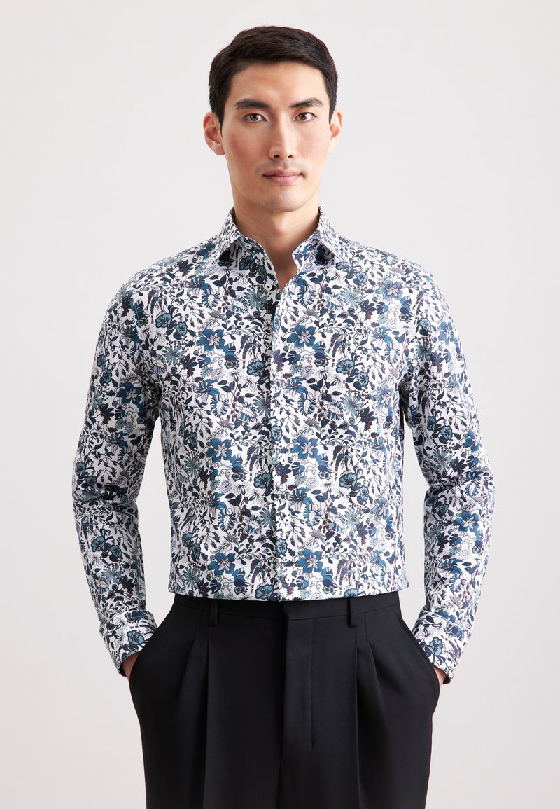 Uomo in camicia bianca a maniche lunghe con stampa floreale blu, pantaloni neri, in piedi con le mani in tasca contro uno sfondo semplice.