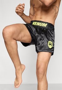 Melni satīna šorti ar elastīgu jostu, uz kuras ir spilgti dzeltens "VENUM" logotips un zaļš akcenta logotips uz kreisās kājas.