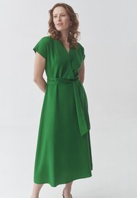 Rochie verde, cu mâneci scurte, talie legată și fustă vaporoasă; realizată dintr-un material neted cu un luciu subtil.