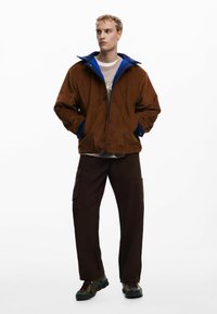Braune Kord-Jacke mit blauer Fütterung, Reißverschluss und zwei vorderen Taschen, kombiniert mit braunen Cargo-Hosen und bunten Turnschuhen.