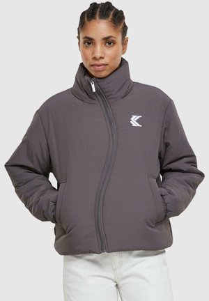 Grijze pufferjack met hoge kraag, voorrits en elastische manchetten. Bevat een wit logo op het bovenste deel van de linkerborst.