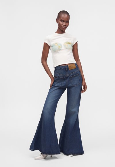 Γυναίκα που φοράει λευκό crop top με εκτυπώσεις παγκόσμιου χάρτη και ψηλόμεσο σκούρο μπλε παντελόνι καμπάνα, στέκεται με το ένα χέρι στην τσέπη.