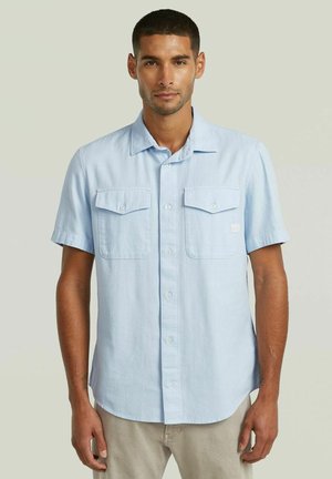 Uomo che indossa una camicia azzurra a maniche corte con due tasche sul petto, in piedi davanti a uno sfondo neutro uniforme.