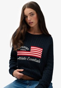 Marineblauwe kabelgebreide trui met een rood-witte gestreepte vlagontwerp en de tekst "Superdry NYC" en "Athletic Essentials" in witte borduursel.