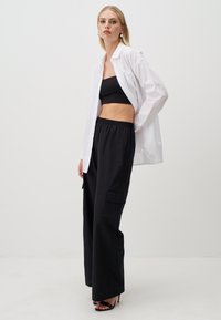Zwarte wijde broek met een textuurstof, gecombineerd met een aansluitend zwart crop top en een witte oversized blouse. Klassieke hoge hakken.