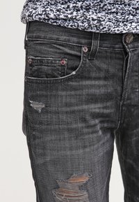 Jeans de mezclilla negra con un diseño desgastado que presenta varios desgarros y hilos sueltos. Incluye detalles de botón de metal y remaches con un acabado envejecido.