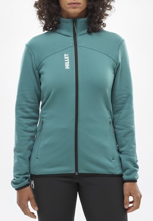 Veste en polaire turquoise zippée avec un col haut, arborant un logo blanc, une fermeture éclair noire et des poches latérales. Associée à un pantalon noir.
