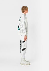 Chaqueta bomber blanca con diseño de mariposa en negro y turquesa, combinada con joggers blancos que presentan gráficos de alas, usados con botas gruesas blancas.