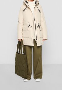 Osoba ubrana w beżową kurtkę z kapturem, oliwkowe szerokie spodnie, białe buty, trzymająca dużą pikowaną torebkę w kolorze oliwkowym, stojąca na szarym podłożu.