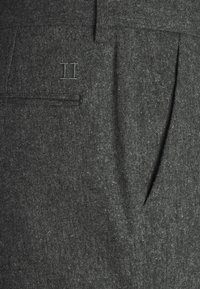 Tissu texturé gris foncé avec un logo double H cousu et une petite poche horizontale sur un vêtement sur mesure.