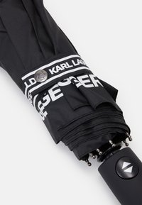 Czarny parasol z gładkim materiałowym nałożeniem, z czarną rączką, metalowym przyciskiem i pasiastą wstążką z białym logo "KARL LAGERFELD".