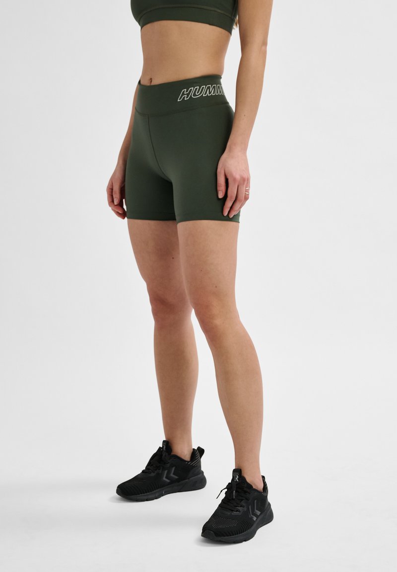 Dunkelgrüne Sportshorts mit hohem Bund, ausgestattet mit einem weißen Logo, aus dehnbarem, glattem Stoff, kombiniert mit schwarzen Turnschuhen.