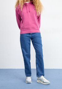 Felpa rosa con logo frontale, abbinata a jeans blu a gamba dritta e sneaker bianche. La felpa presenta un cappuccio con cordino e polsini a coste.